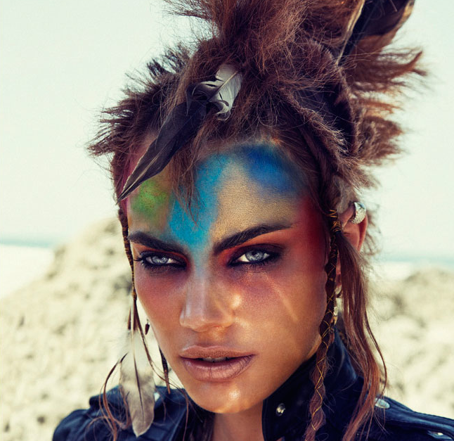 A woman with colorful makeup and feathers posing in the sand.