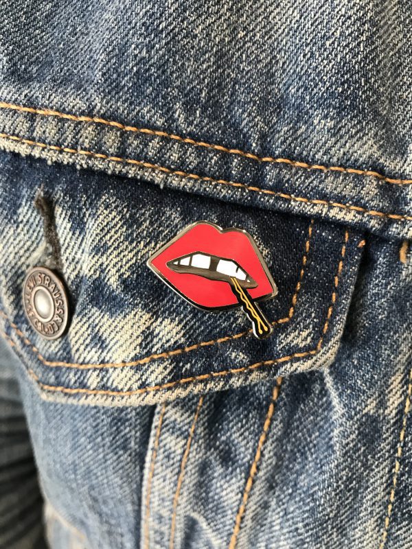 A "Lips" enamel lapel pin by LH™ with a red lipstick on it in the pocket of someone's jeans.