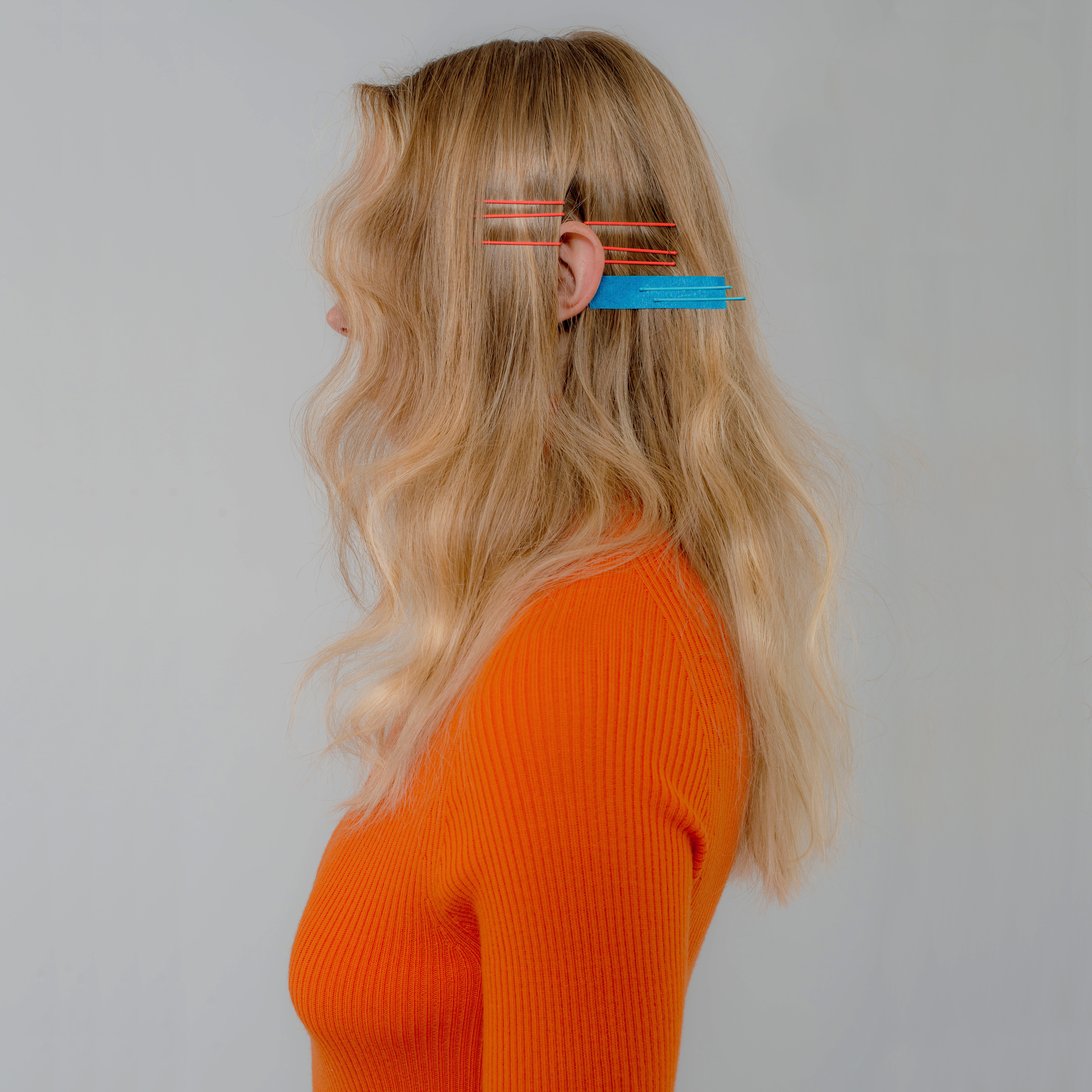 Profile of blonde woman wearing orange sweater with blue and red hair clips.